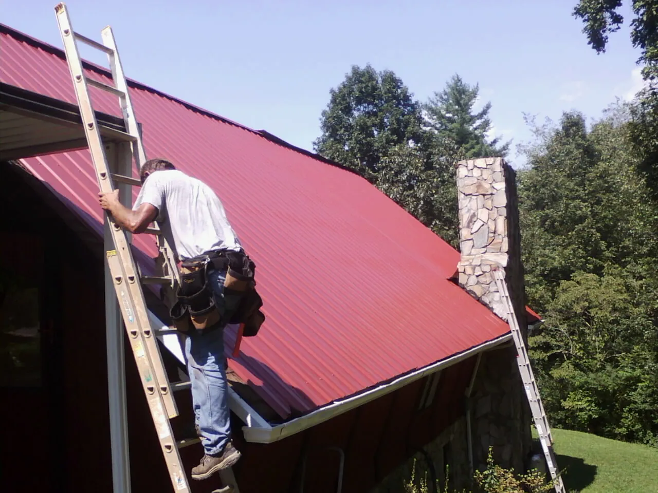 Roofer on a ladder beside a red metal roof and stone chimney — Soffit & Fascia Repair by Roofing Brothers in Padroni